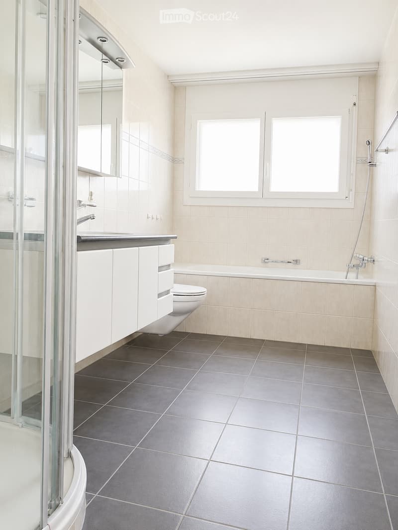 Modern bathroom with white tiles, dark grey floor tiles, a bathtub, toilet, sink, and a mirrored cabinet.