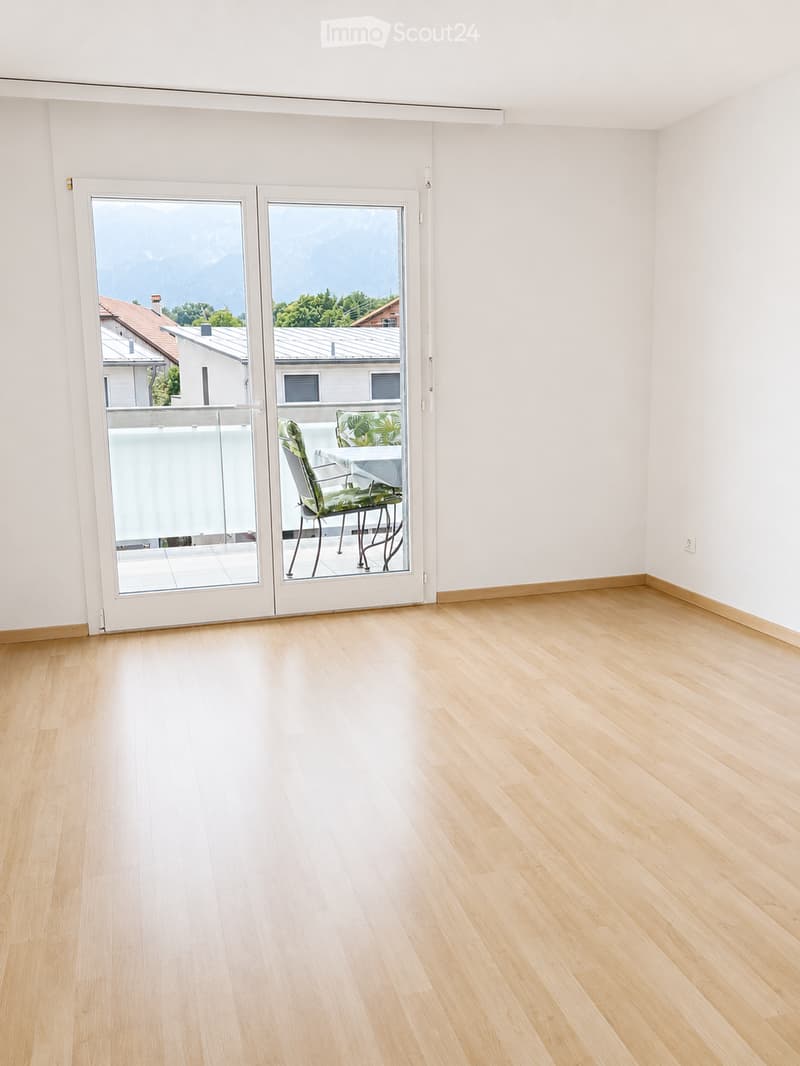 Empty room with light wooden flooring and white walls, featuring a large sliding glass door leading to a balcony with outdoor furniture visible.