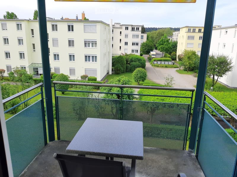 Balcony with a table and chair, overlooking a residential area with buildings and greenery.