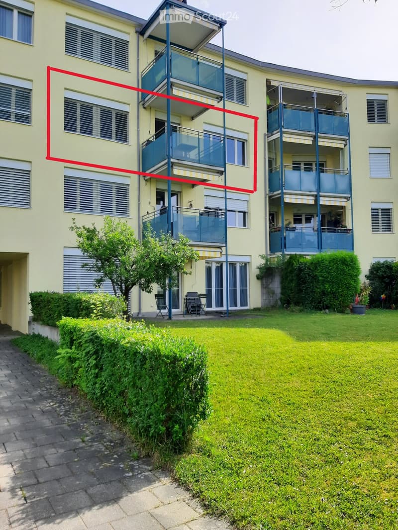 Multi-story residential building with light yellow facade, multiple balconies, and surrounding greenery.