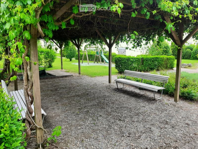 A shaded outdoor area with gravel ground, wooden benches, and a playground in the background.