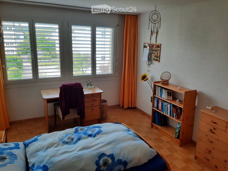 Cozy bedroom with wooden flooring, large windows with blinds, a bed with blue floral bedding, a wooden desk with a chair, a bookshelf filled with books, a small drawer unit, and decorative items including a dreamcatcher and a flower vase.