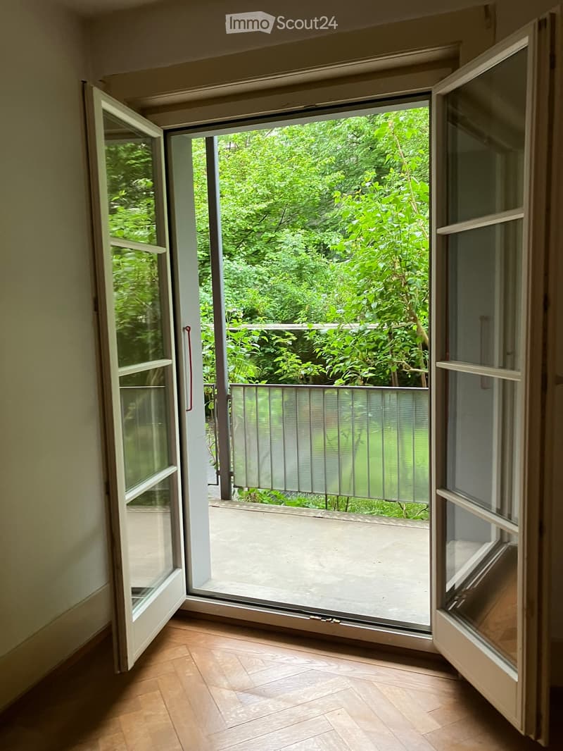 Open sliding glass door leading to a balcony with a metal railing, surrounded by lush green foliage.