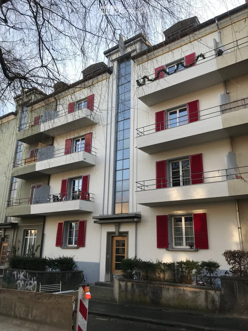 Building. Multi-story residential building with white facade, red shutters, and multiple balconies. Features a mix of modern and traditional architectural elements.