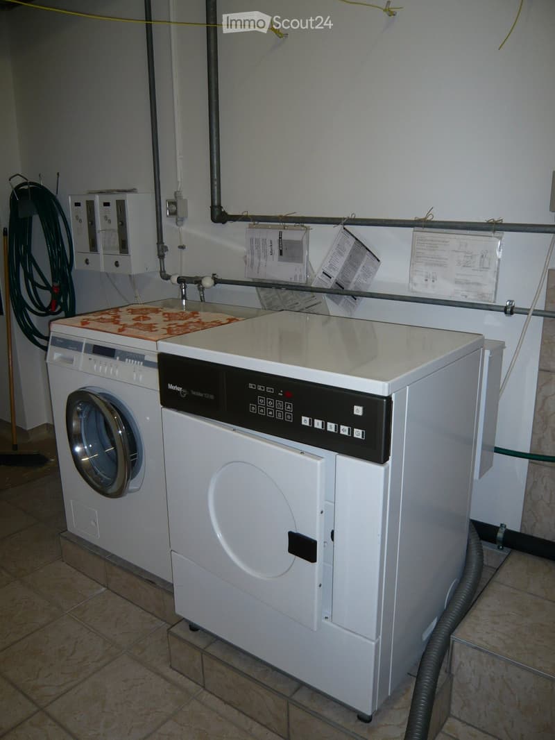A small utility room with a washing machine and dryer, tiled floor, and various pipes and hoses.