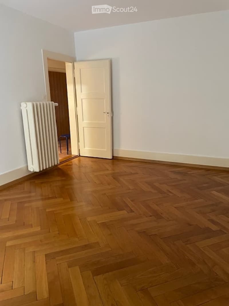 Empty room with wooden herringbone flooring, white walls, and a white radiator. A door is open to an adjacent room.