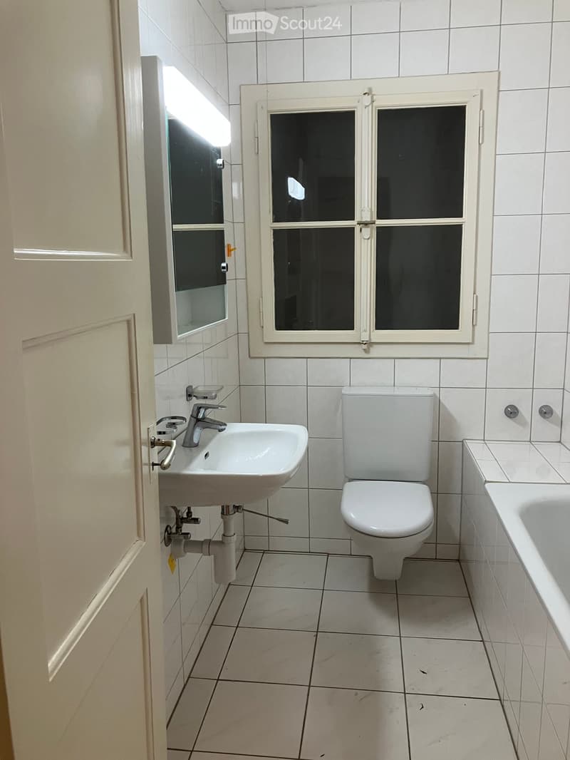 Bathroom with white tiles, a sink with a faucet, a toilet, a bathtub, and a mirror above the sink. A window with frosted glass is visible on the wall.