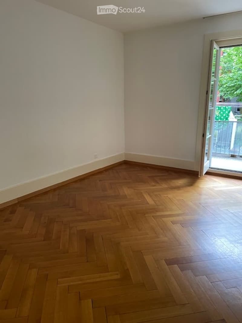 Empty room with wooden herringbone flooring, white walls, and an open door leading to a balcony.