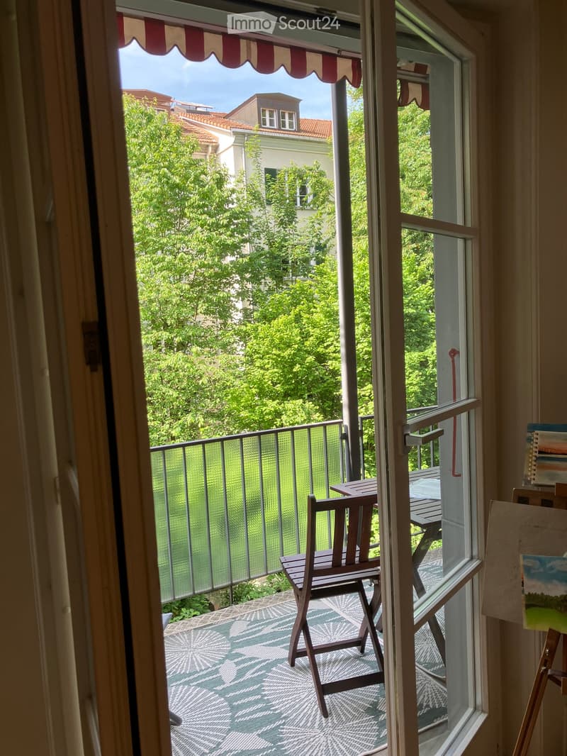 Balcony with a wooden chair and table, overlooking a green garden area with trees and neighboring buildings.