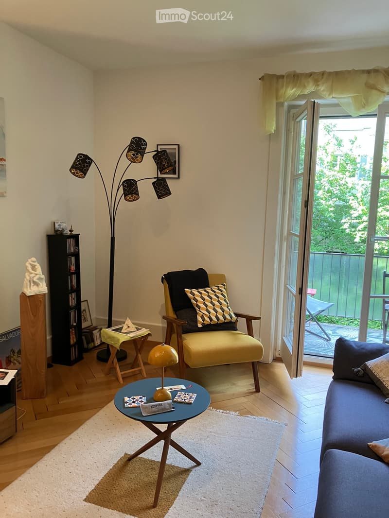 Living room with a blue sofa, yellow armchair, small round table, floor lamp, bookshelf, and a view to a balcony.