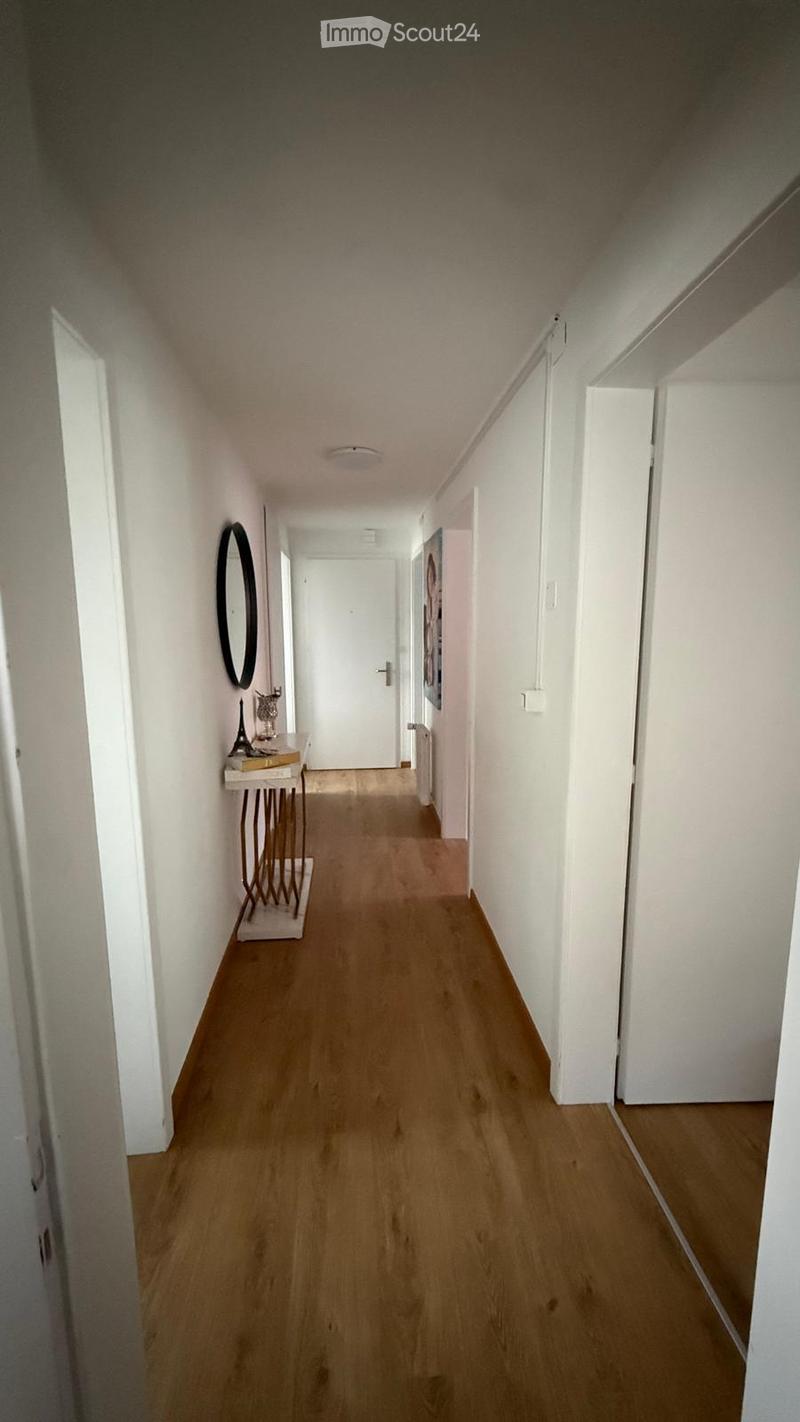 A narrow hallway with wooden flooring, white walls, and a small table with a mirror and decorative items on the left side.