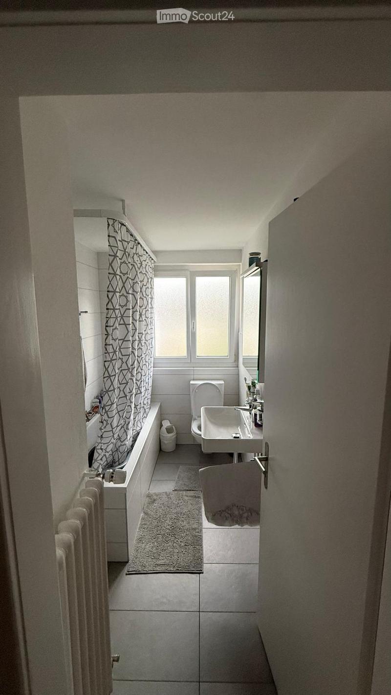 Modern bathroom with a bathtub, toilet, sink, radiator, mirror, towel, and a patterned shower curtain. The space is well-lit with natural light from a window and features tiled floors and walls.