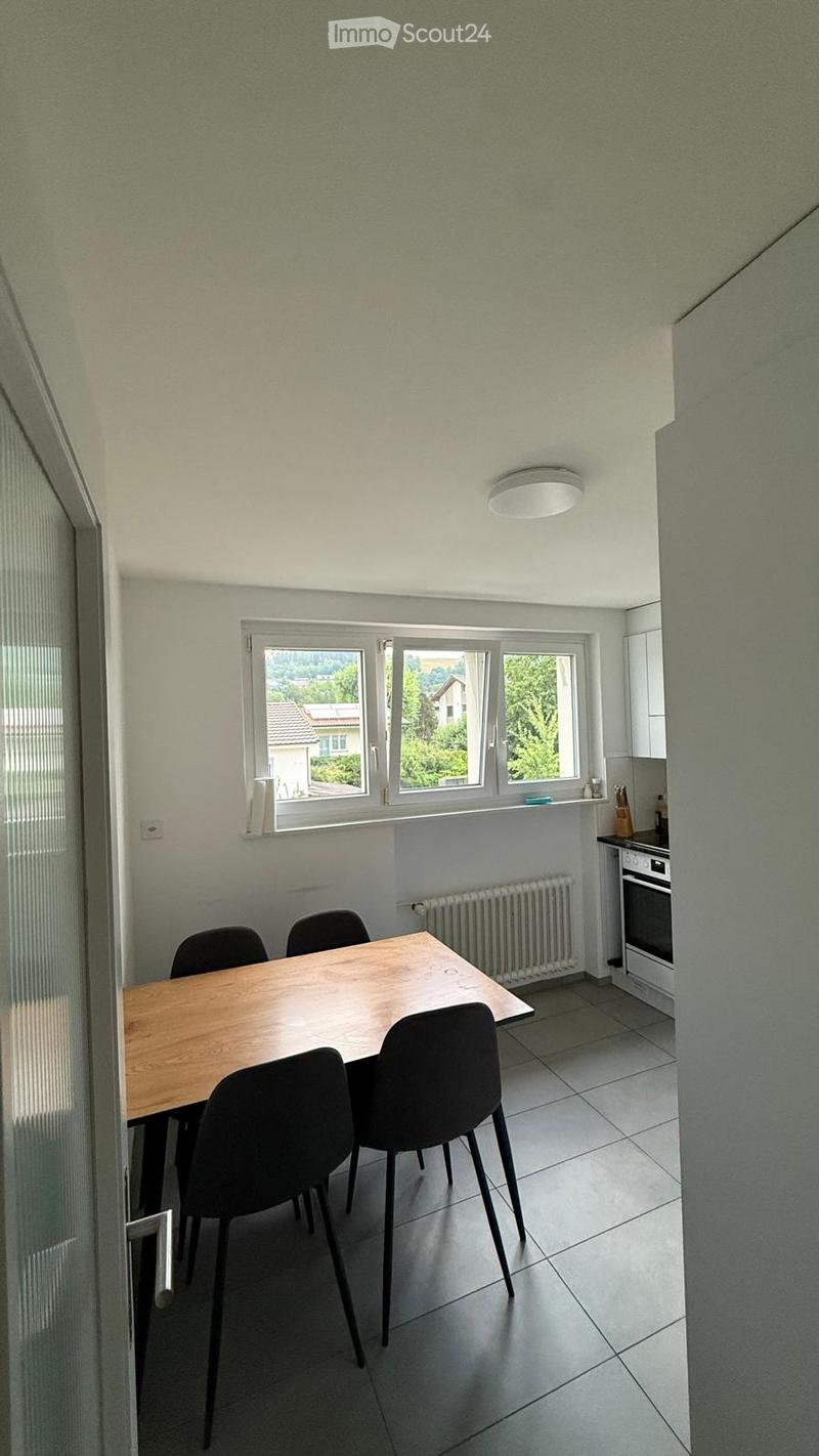 A small, modern kitchen-dining area with a wooden table, four black chairs, a radiator, and an oven. The room has large windows offering a view of greenery and houses outside.