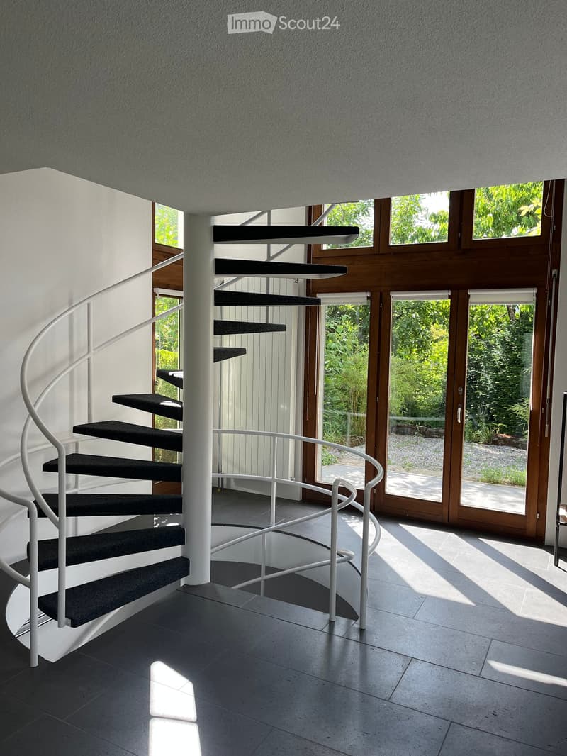Spiral staircase with black steps and white railing, leading to an open area with large windows and a view of greenery outside.