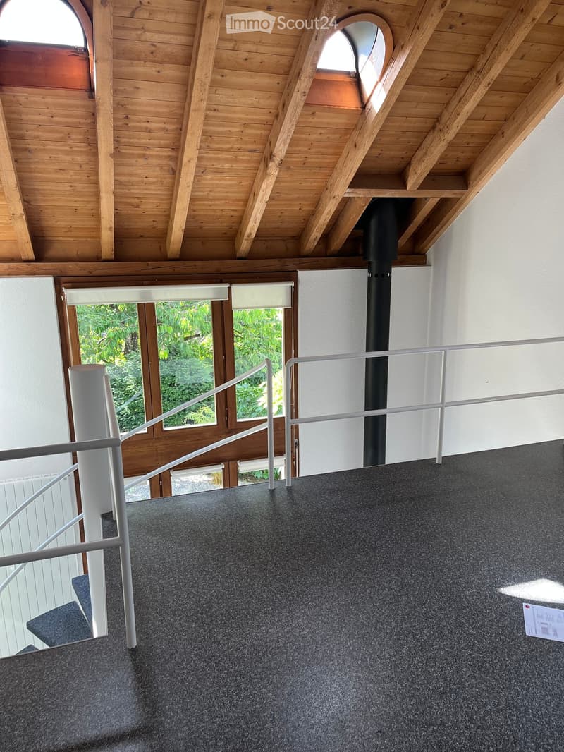 Modern interior staircase with wooden ceiling beams, grey carpeted steps, white metal railing, and large windows overlooking greenery.