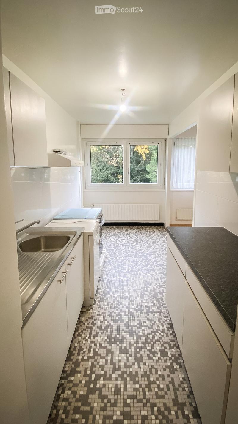 Modern kitchen with white cabinets, dark countertop, and a tiled floor. Features a sink, window with a view of greenery, and a radiator below the window.