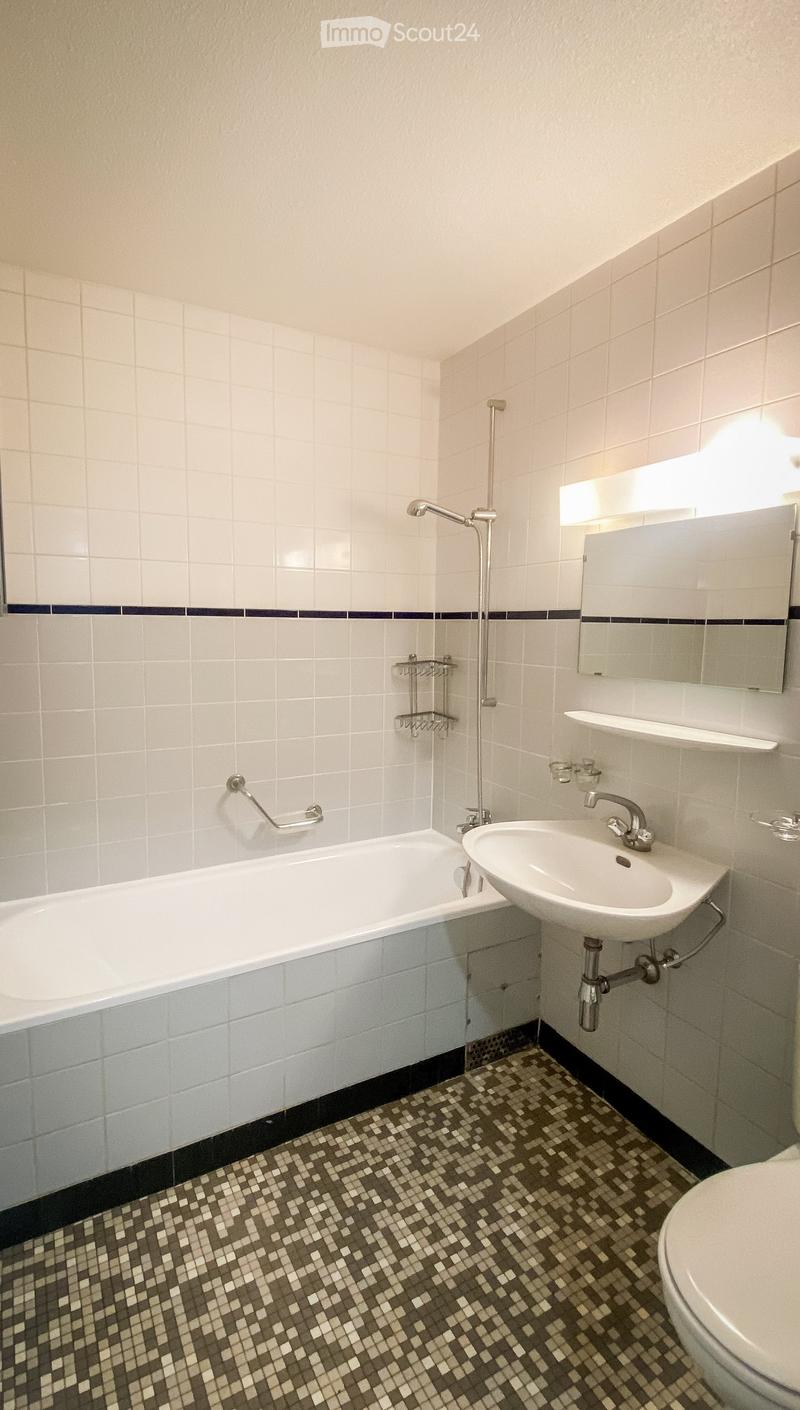 Modern bathroom with white tiled walls, a bathtub, a sink, a toilet, a mirror, and a shower handle. The floor features a patterned tile design.