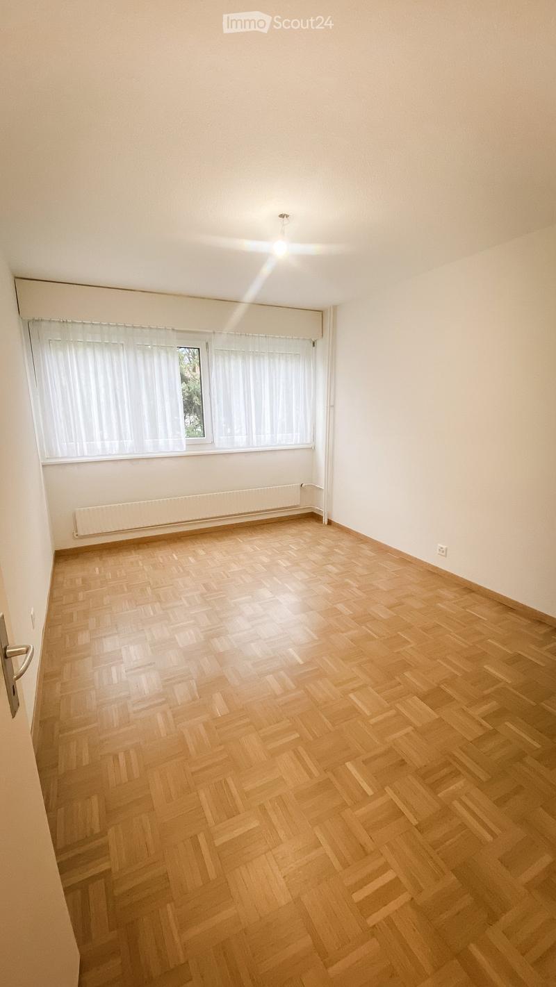 Empty room with wooden herringbone flooring, white walls, large window with white curtains, radiator, and a door with a silver handle.