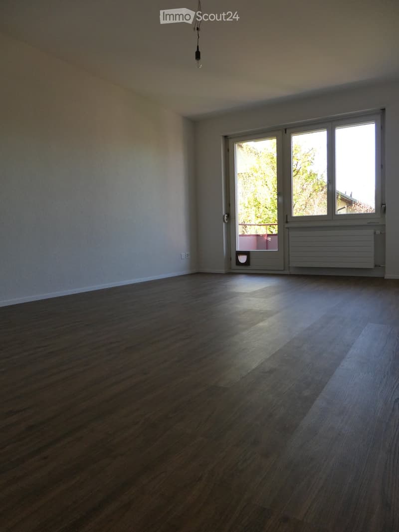 Empty room with wooden flooring, white walls, and large windows with a sliding door leading to a balcony.