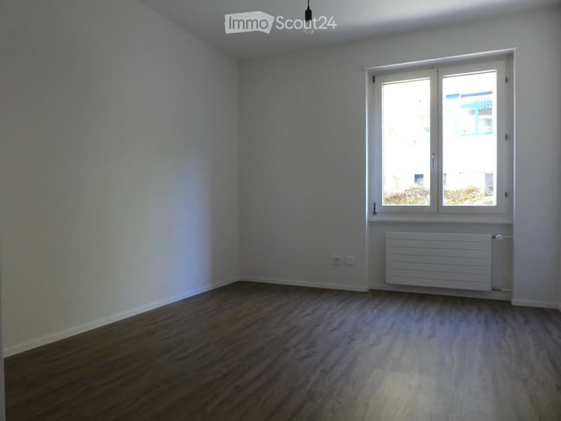 Empty room with light-colored wooden flooring, white walls, and a large window with white frame. A white radiator is placed below the window.
