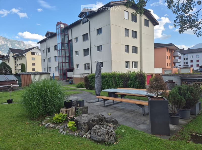 Well-maintained garden area with a wooden table and bench, surrounded by greenery, potted plants, and decorative rocks.