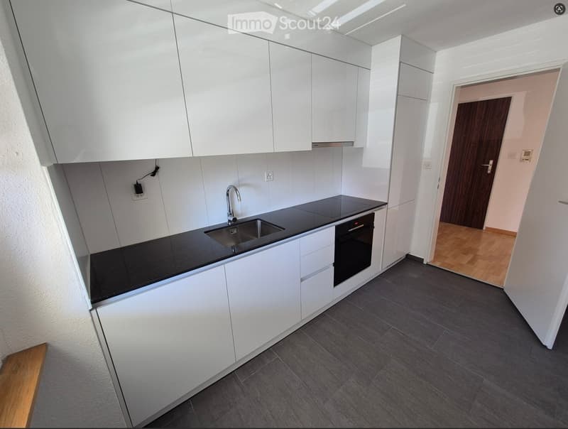 Modern kitchen with white cabinets, black countertop, stainless steel sink, and built-in oven. Grey tiled floor and wooden flooring visible in adjacent area.