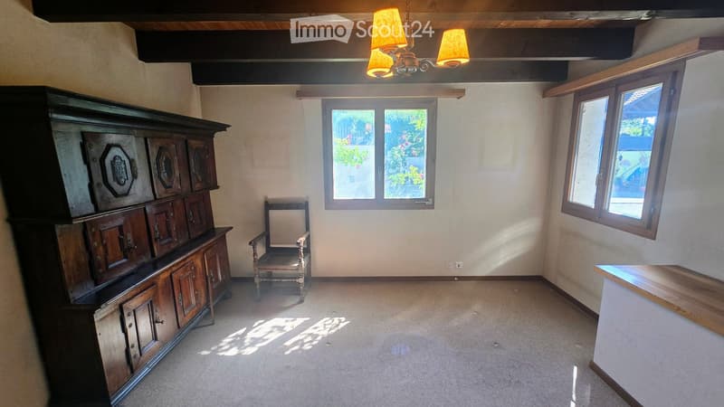A furnished room with a large wooden cabinet, a wooden chair, and large windows letting in natural light. The room has a carpeted floor and exposed wooden beams on the ceiling.