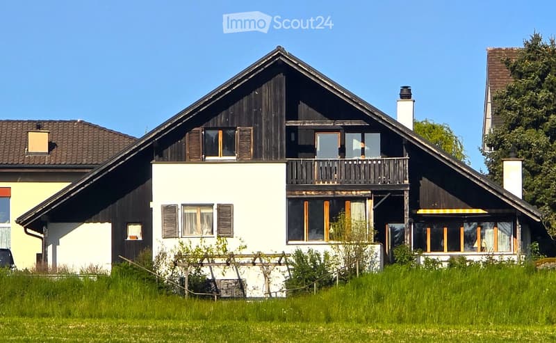 House. Two-story house with a mix of white and dark wood exterior, featuring a balcony on the upper level, multiple windows with shutters, and a well-maintained lawn in front.