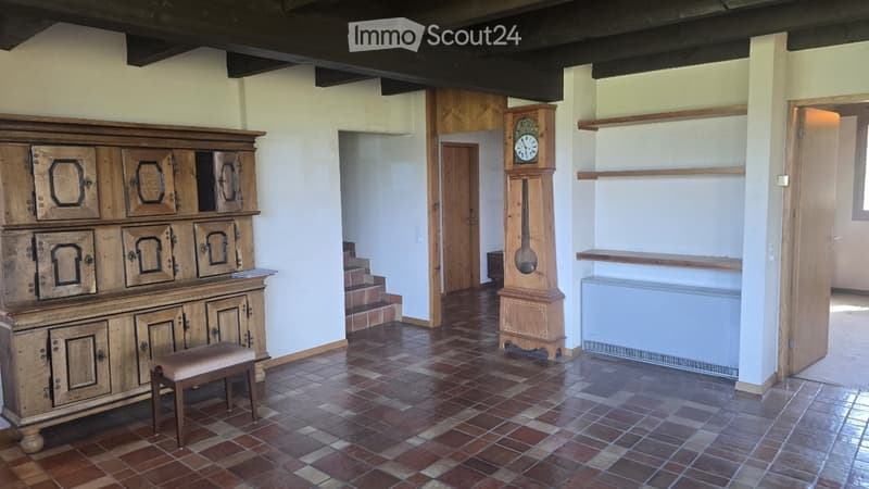 A spacious, well-lit room with exposed wooden beams, a large wooden cabinet, a grandfather clock, a small wooden stool, and built-in wooden shelves. The floor is tiled in a warm, earthy pattern.