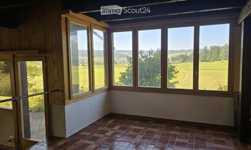 Bright, empty room with large windows offering a scenic countryside view, wooden walls, and a tiled floor.