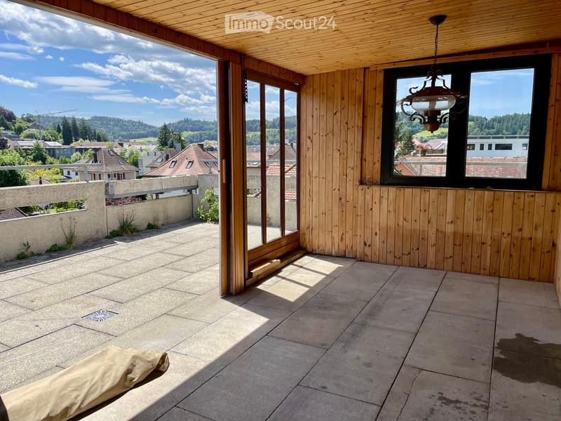 Spacious balcony with wooden walls and ceiling, large windows offering scenic views, tiled floor, and a single piece of furniture.