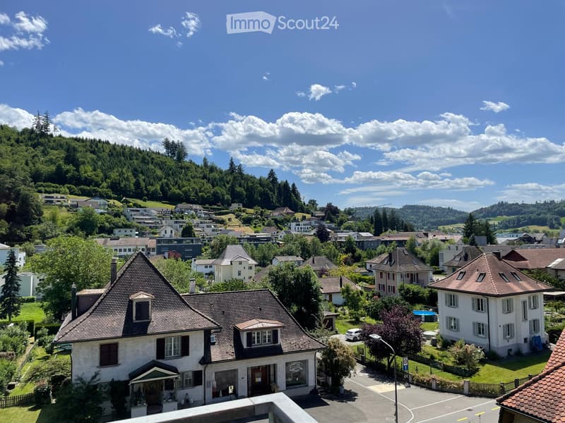 A scenic view of a residential area with multiple houses, surrounded by lush green hills and trees under a partly cloudy sky.