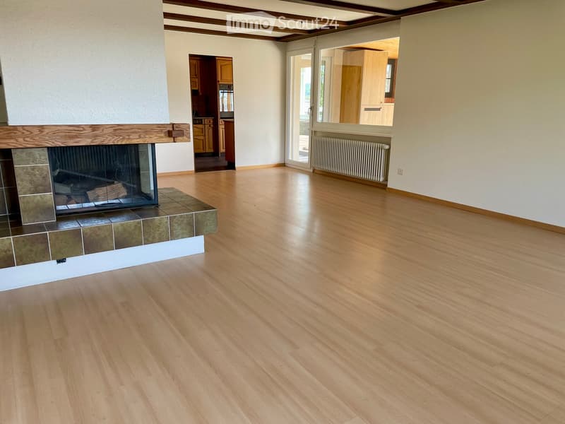 Empty room with wooden flooring, fireplace, and large windows leading to another room.