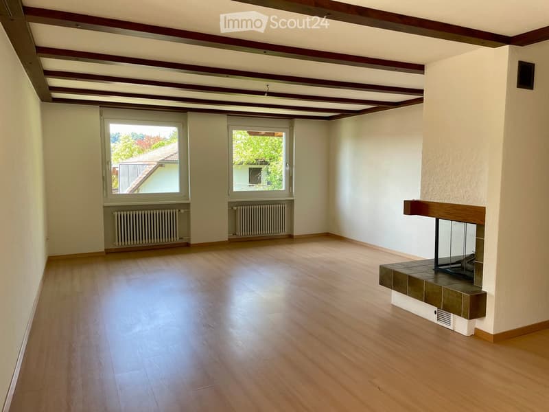Empty room with wooden flooring, white walls, exposed wooden ceiling beams, fireplace, and large windows with radiators beneath them.