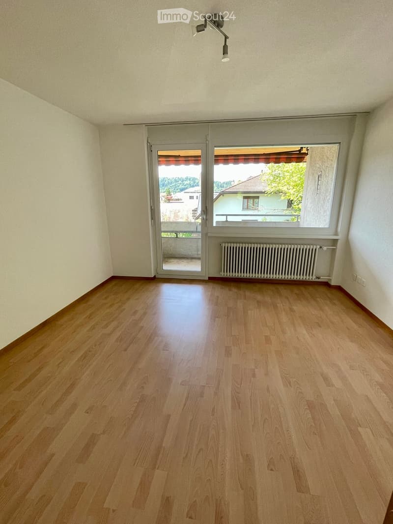 Empty room with wooden flooring, large window, sliding glass door leading to a balcony, radiator, and ceiling light fixture.