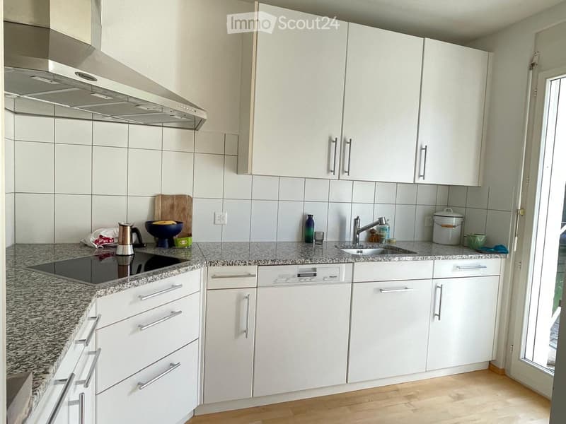 Modern kitchen with white cabinets, granite countertops, and stainless steel appliances including a dishwasher and stove. Features a sink with a modern faucet, various kitchen items on the counter, and a wooden cutting board.