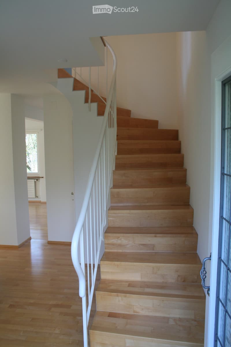 Spiral staircase with wooden steps and white railing, leading to an open area with wooden flooring and white walls.