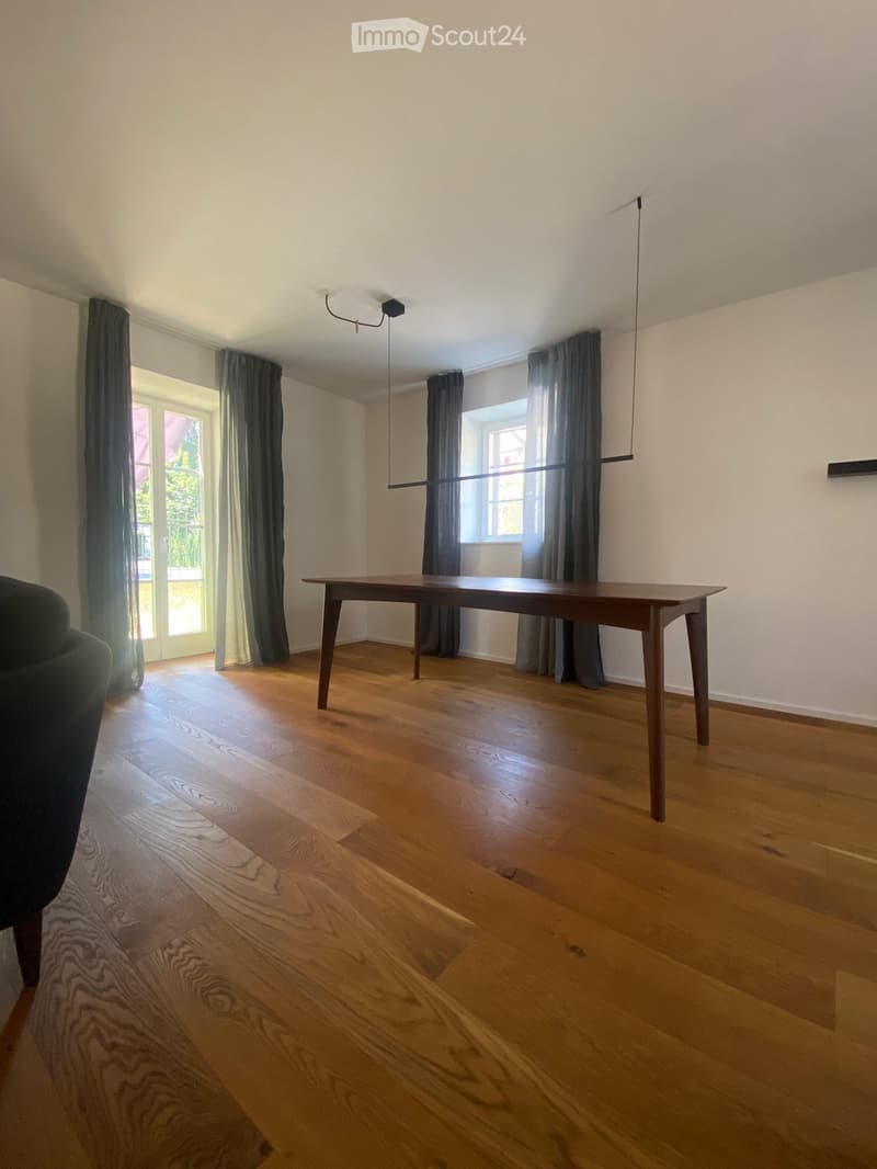 Bright, unfurnished room with wooden flooring, large windows, and a single table and sofa.