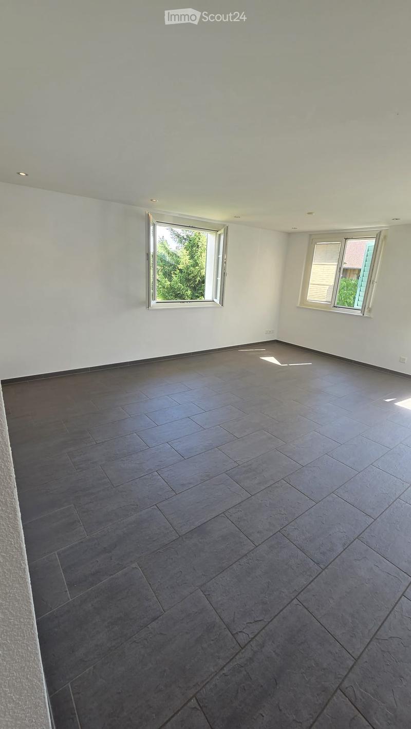 Empty room with grey tiled floor, white walls, and two windows, one of which is open to show greenery outside.