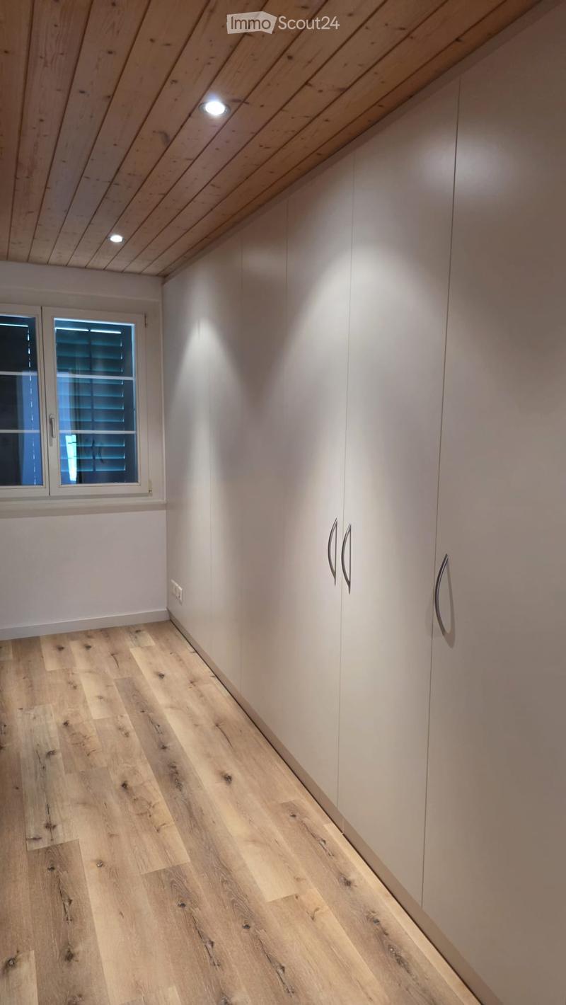 A narrow hallway with light wooden flooring, white walls, built-in white cabinets with silver handles, and a small window with blinds on the left side.