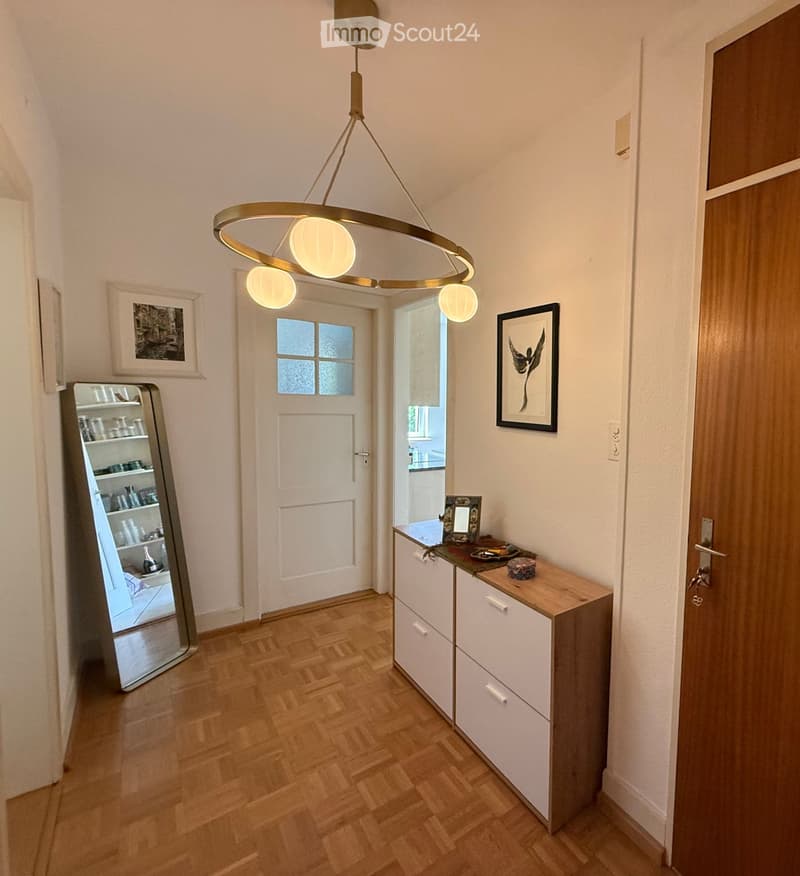 Hallway with wooden flooring, white walls, a large round pendant light fixture, a small console table with drawers, and a mirror reflecting a kitchen area.