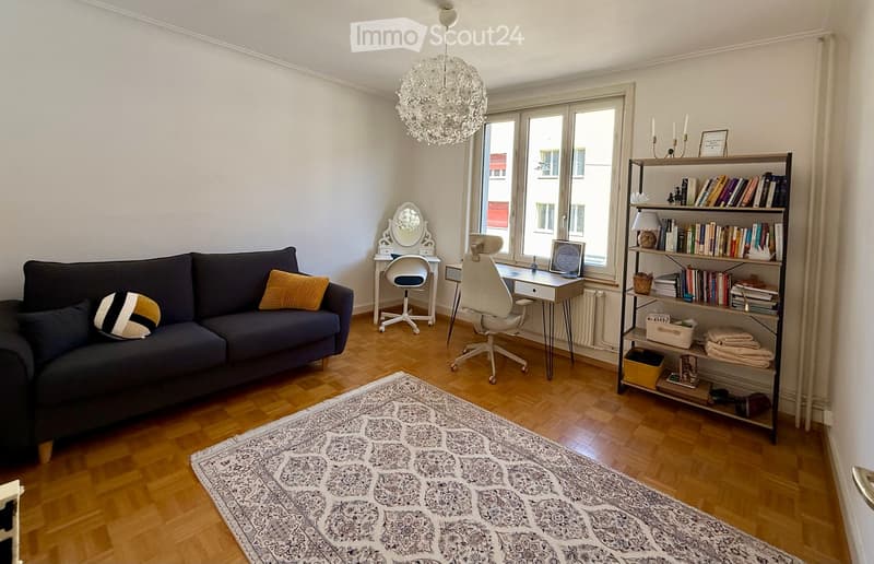 A well-lit living room with a dark blue sofa, a patterned rug, a bookshelf filled with books, a desk with a laptop, and a vanity with a mirror.