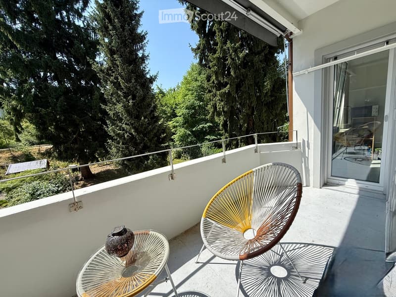 Balcony with modern chairs and a small table, overlooking a green area with trees.