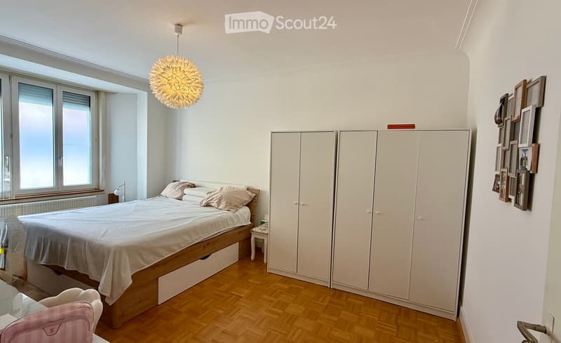 Bright bedroom with wooden flooring, large windows, a double bed with pillows and blankets, a wardrobe, a small stool, and a pink chair. A decorative light fixture hangs from the ceiling, and picture frames are displayed on the wall.