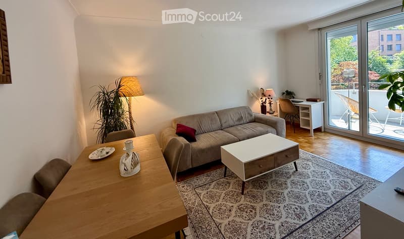 Bright living room with a grey sofa, coffee table, dining table, and a desk area near a large sliding glass door leading to a balcony.