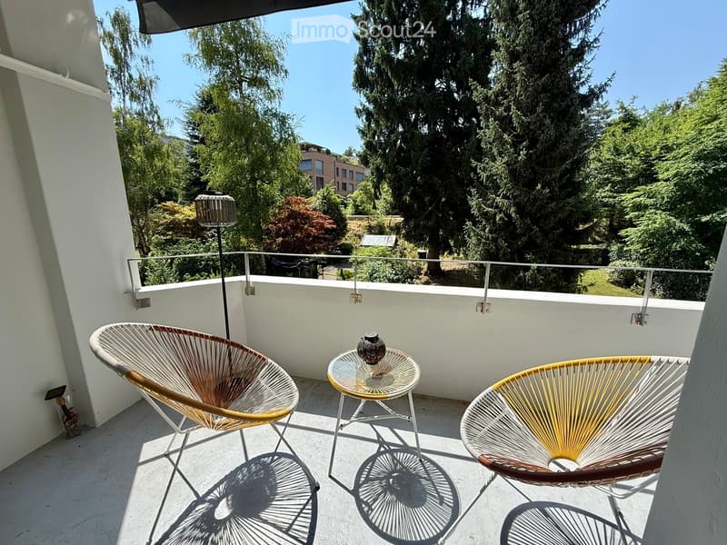 Balcony with two modern chairs, a small round glass table, and a decorative vase, overlooking a lush green area with trees.