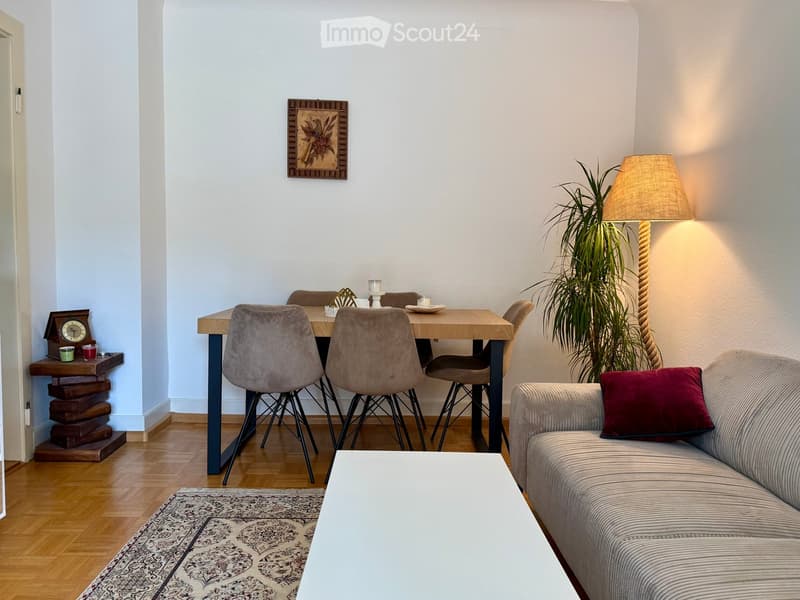 A modern living room with a sofa, coffee table, dining table, chairs, a floor lamp, a plant, a clock, and a picture frame on the wall.