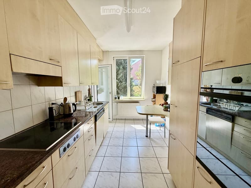 Bright kitchen with light wood cabinets, tiled floor, and modern appliances including a stove, oven, sink, and dishwasher. A small table and chair are placed near the window, which offers a view of greenery outside.