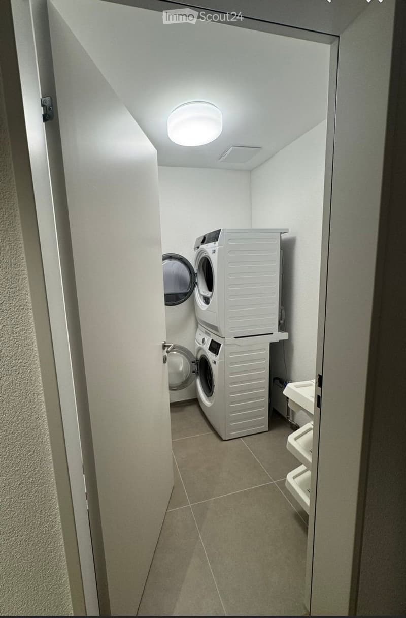 A small, well-lit laundry room with a stacked washer and dryer unit, a small sink, and tiled flooring.