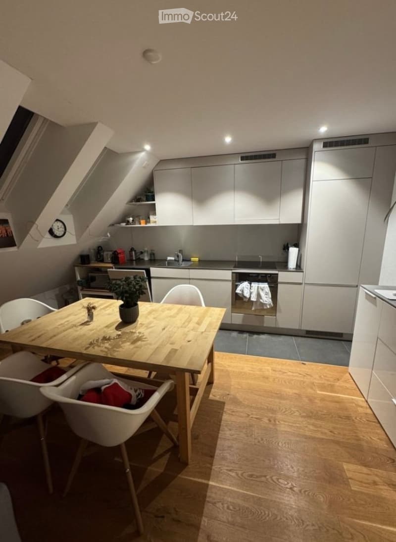 Modern kitchen with light grey cabinets, wooden dining table, white chairs, and a small potted plant on the table. The kitchen features an oven, microwave, and refrigerator, with a window and a clock visible on the wall.